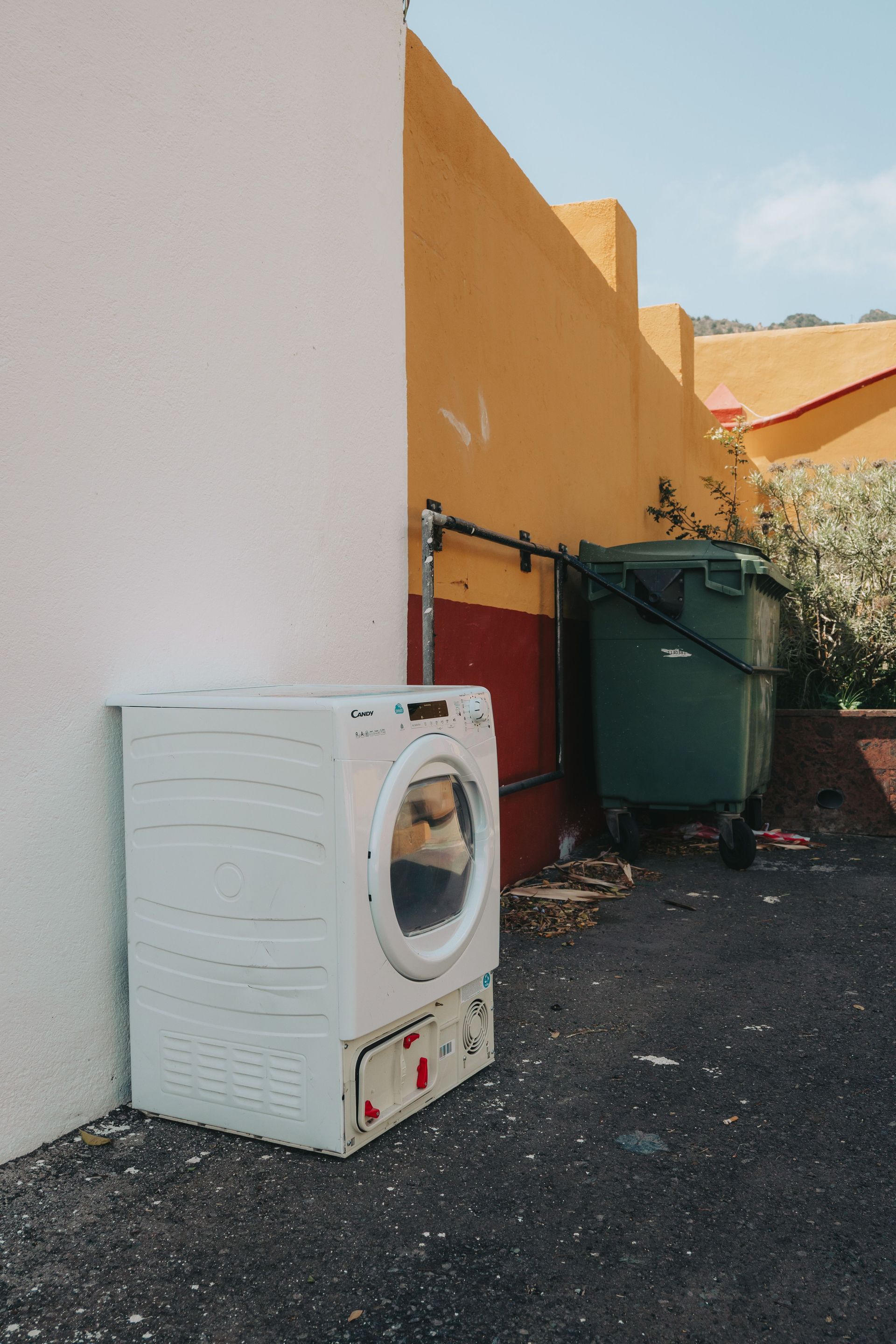 A discarded washing machine by a green trash bin on a vibrant street in Vallehermoso.
